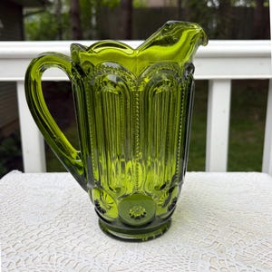 May include: A green glass jug with a handle and a spout. The jug features a textured, patterned design with vertical lines and starburst details. The jug is placed on a white lace tablecloth.