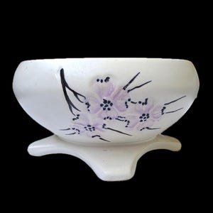 以下が含まれることがあります： 三本足の白い陶器のボウル。ボウルは手描きの紫色の花と黒い茎で装飾されています。ボウルは丸みを帯びた形で、装飾目的にも適しています。