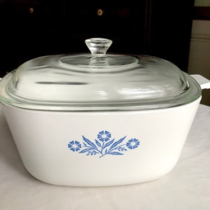 Könnte beinhalten: Weißer Auflaufform mit einem Deckel und Knopf aus klarem Glas. Die Auflaufform hat ein blaues Blumendesign. Sie hat weiße Griffe auf jeder Seite.
