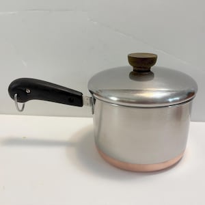 May include: A stainless steel saucepan with a copper-colored base and a matching lid. The pan features a black handle with a small metal hanging loop attached to the end. The lid has a dark brown, circular knob centered on the top surface.