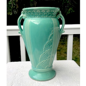 Puede incluir: Un jarrón de cerámica azul turquesa con dos asas y un patrón floral decorativo. El jarrón tiene un borde festoneado y una base ancha.