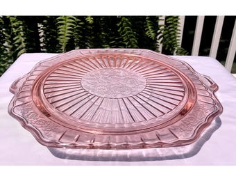 Puede incluir: Una fuente para pastel de vidrio rosa con borde festoneado. La fuente presenta un diseño floral central y líneas radiales. La fuente es de vidrio y de color rosa claro. La fuente es redonda y tiene un diámetro de aproximadamente 30 cm.