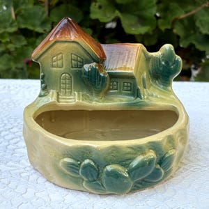 May include: A ceramic house-shaped planter with a brown roof and green walls. The planter has a rectangular opening and is surrounded by green foliage. The base is a light beige colour.