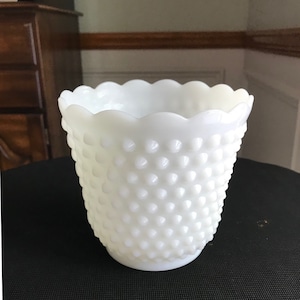 Könnte beinhalten: Eine weiße Hobnail-Vase mit einem gewellten Rand. Die Vase hat eine strukturierte Oberfläche mit kleinen, abgerundeten Erhebungen, die die Außenseite bedecken. Die Vase steht auf einer schwarzen Oberfläche.