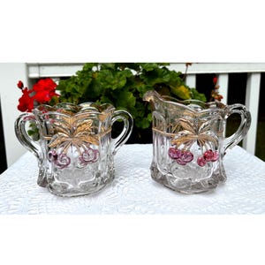 Puede incluir: Un juego de azucarero y jarra de crema de cristal transparente. El juego presenta un diseño de hojas doradas y cerezas. El azucarero tiene dos asas y la jarra de crema tiene un pico. El juego está sobre una superficie blanca con vegetación en el fondo.