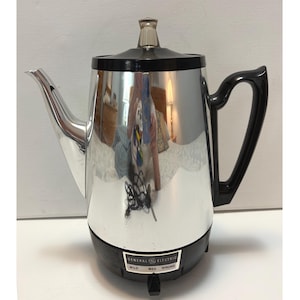 Może przedstawiać: Zabytkowy elektryczny ekspres do kawy General Electric z błyszczącym chromowanym wykończeniem. Ma czarną podstawę, uchwyt i pokrywę, z przezroczystym szklanym pokrętłem. Panel sterowania wskazuje "Mild, Med, Strong".