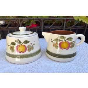 Puede incluir: Un azucarero vintage con tapa y una jarra de crema. Ambas piezas son blancas con una franja verde y presentan un diseño pintado a mano de una manzana amarilla con detalles rosas y hojas verdes. La jarra de crema tiene un interior marrón.