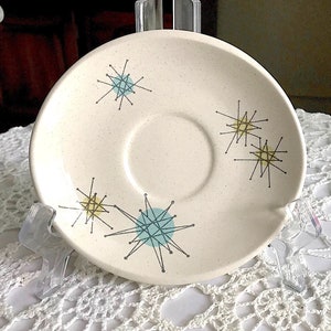 May include: A white saucer with a speckled glaze features a mid-century modern design of blue, yellow, and black starbursts.