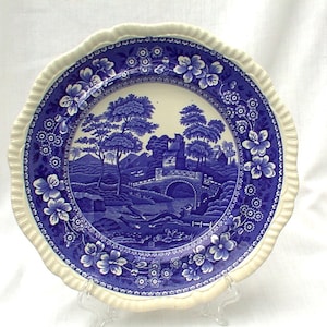 Può includere: Un piatto decorativo blu e bianco con una scena paesaggistica. Il piatto presenta un'illustrazione dettagliata di un ponte, alberi e un castello. Il bordo è smerlato e decorato con motivi floreali.
