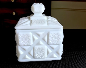 May include: A white milk glass box with a lid. The box has a square shape and is decorated with a geometric pattern. The lid has a knob on top.