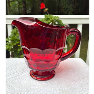 May include: A ruby red glass jug with a handle. The jug has a faceted design on the lower portion and a spout for pouring. The jug is sitting on a white surface.