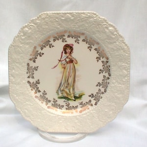 May include: A white square ceramic plate with a raised embossed design around the edge. The centre of the plate features a gold-rimmed circle with a floral design and a painted image of a young girl in a white dress and pink hat.