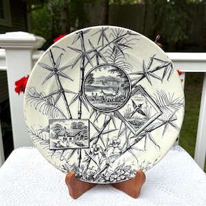 May include: Cream-coloured decorative plate with black bamboo and scenic illustrations. The plate features three framed scenes: a deer in a landscape, a bird in flight, and a cottage. The plate is displayed on a wooden stand.