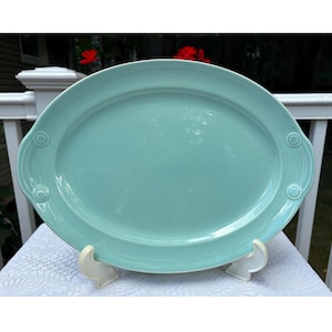 May include: A large, oval serving platter in a soft turquoise colour. The platter has decorative detailing on the ends and is supported by a white stand. The smooth surface is ideal for serving food.