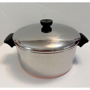 May include: A stainless steel cooking pot with a polished silver finish and a matching lid. The pot features two black side handles and a black knob on the centre of the lid. The base of the pot has a visible copper-coloured band around the bottom edge.