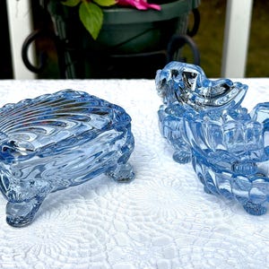 May include: Two light blue pressed glass trinket dishes with a shell-inspired design. One dish features a rectangular shape with a lid, while the other is an open bowl. Both pieces are supported by four small feet and have a scalloped edge pattern.