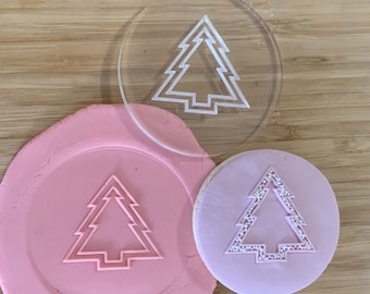 Pode incluir: Um carimbo de biscoito de acrílico transparente com um desenho de árvore de Natal, junto com uma impressão de argila rosa e um biscoito decorado. O biscoito tem cobertura rosa claro com um desenho de árvore e granulado. O carimbo é redondo.
