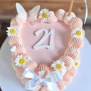 May include: A heart-shaped pink cake decorated with white daisies, a white butterfly, and a white bow. The number "21" is displayed in the centre. A single candle with a white ribbon is placed on top. The cake sits on a white plate.