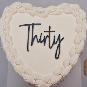 Op de afbeelding: Witte hartvormige taart met witte frosting en een zwarte inscriptie met de tekst "Thirty".