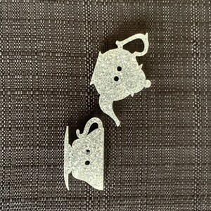 May include: Two white glitter teacup and teapot ornaments. The teapot has a spout and handle. The teacup has a handle and a saucer.