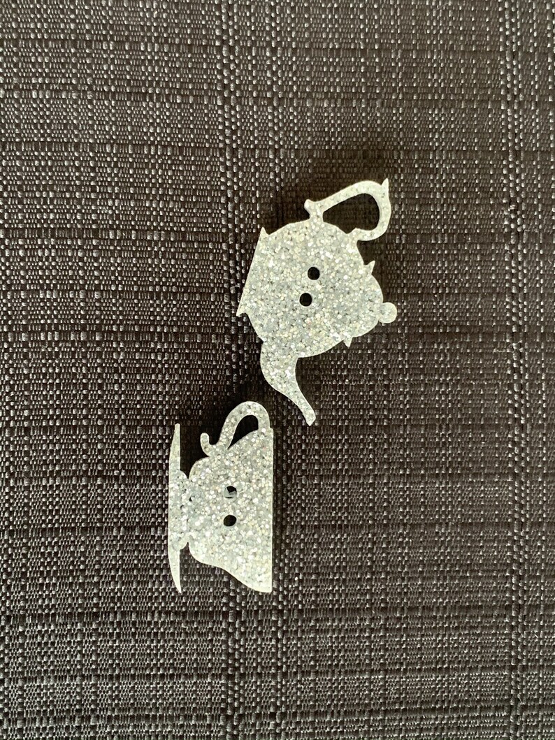 May include: Two white glitter teacup and teapot ornaments. The teapot has a spout and handle. The teacup has a handle and a saucer.