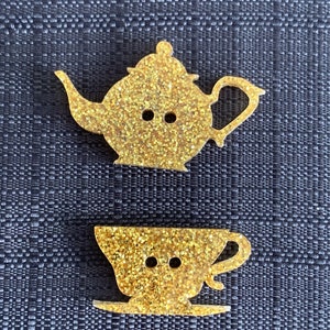 May include: Two gold glitter buttons shaped like a teapot and a teacup. The teapot has a spout and a handle. The teacup has a handle and a saucer.