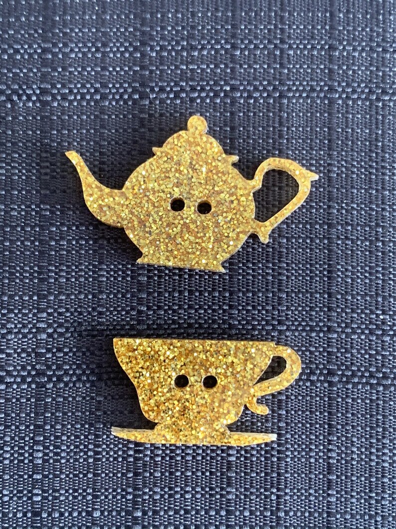May include: Two gold glitter buttons shaped like a teapot and a teacup. The teapot has a spout and a handle. The teacup has a handle and a saucer.