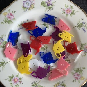 May include: A collection of colorful plastic teapots in various colors, including pink, yellow, blue, red, purple, and white, on a floral patterned plate.