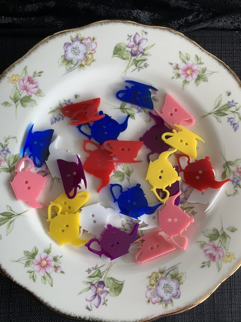 May include: A collection of colorful plastic teapots in various colors, including pink, yellow, blue, red, purple, and white, on a floral patterned plate.