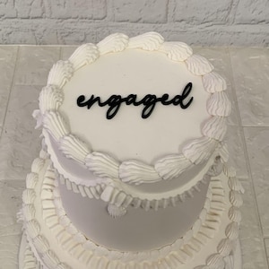 May include: A multi-tiered white cake with white frosting and decorative piping. The top layer has the word "engaged" in black script. This cake is a celebratory dessert, ideal for an engagement party.
