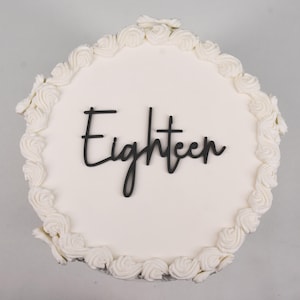 May include: A round white cake decorated with white frosting and a border of piped buttercream swirls. The word "Eighteen" is written in black script across the top of the cake. A celebratory dessert.