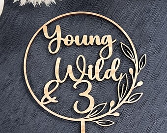 May include: Wooden cake topper with a circle design and the words "Young Wild & 3" with leafy accents.