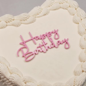May include: A white heart-shaped cake with white frosting and a pink "Happy Birthday" topper.