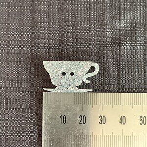 May include: A white glitter teacup and saucer button, measuring approximately 2 cm in diameter. The button is shaped like a teacup with a handle and a saucer.