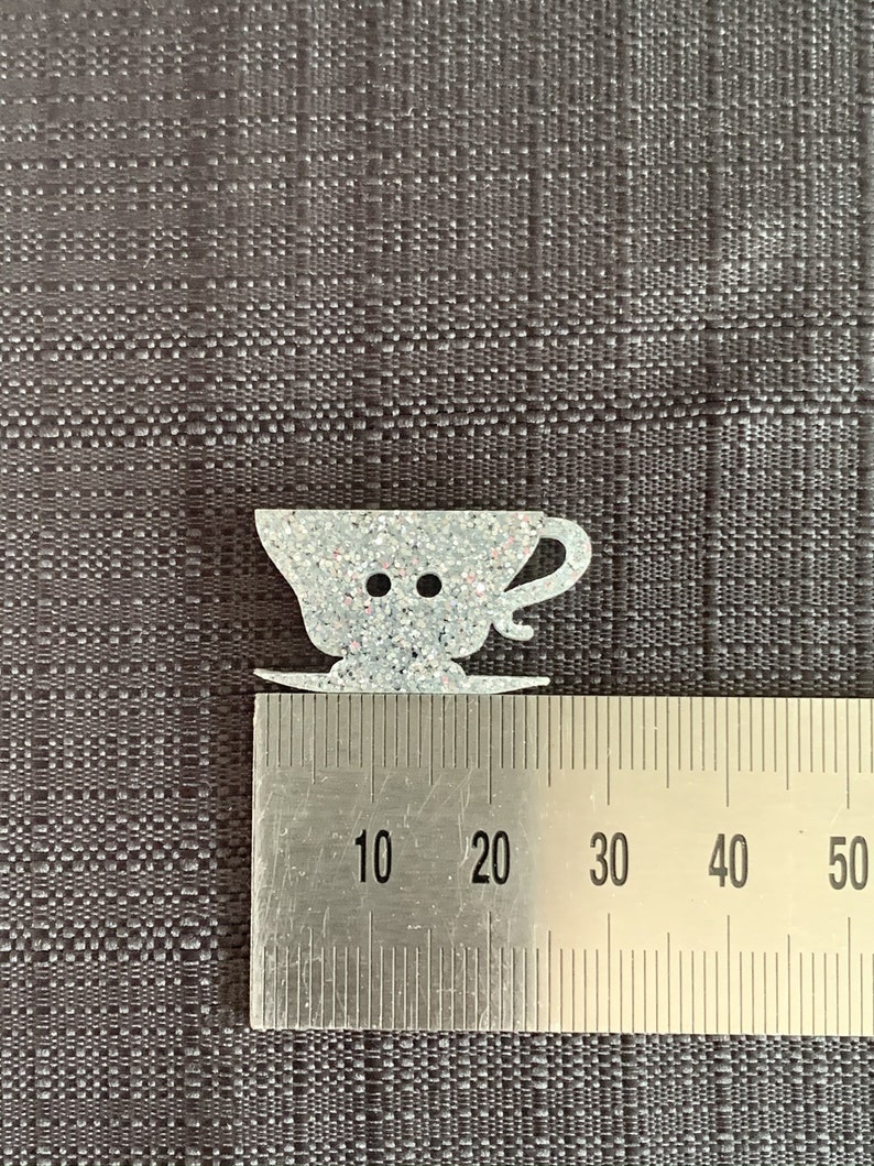 May include: A white glitter teacup and saucer button, measuring approximately 2 cm in diameter. The button is shaped like a teacup with a handle and a saucer.