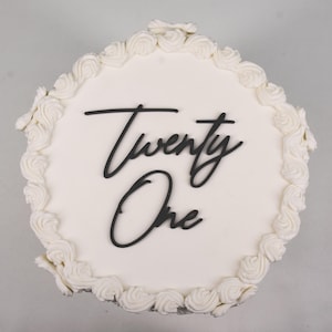 Könnte beinhalten: Ein runder weißer Kuchen mit einem dekorativen Rand aus weißen Zuckergusswirbeln. Die Worte "Twenty One" sind in schwarzer Schrift über dem Kuchen geschrieben. Ein festliches Dessert.