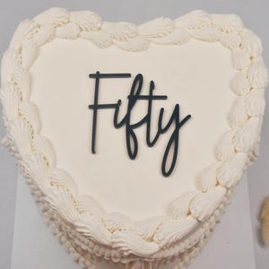 Può includere: Una torta bianca a forma di cuore con glassa bianca e un topper nero 'Cinquanta'.