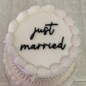 May include: White wedding cake with white icing and a black 'just married' topper.