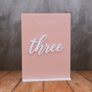May include: A pink acrylic table number with a white stand. The number "three" is written in white cursive lettering on the front of the sign. The text "table" is written in white lettering at the top of the sign.