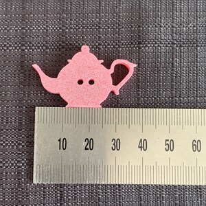 May include: A pink felt teapot cutout measuring approximately 2.5 inches wide. The teapot has a handle and a spout. The cutout is on a gray textured surface with a ruler for scale.