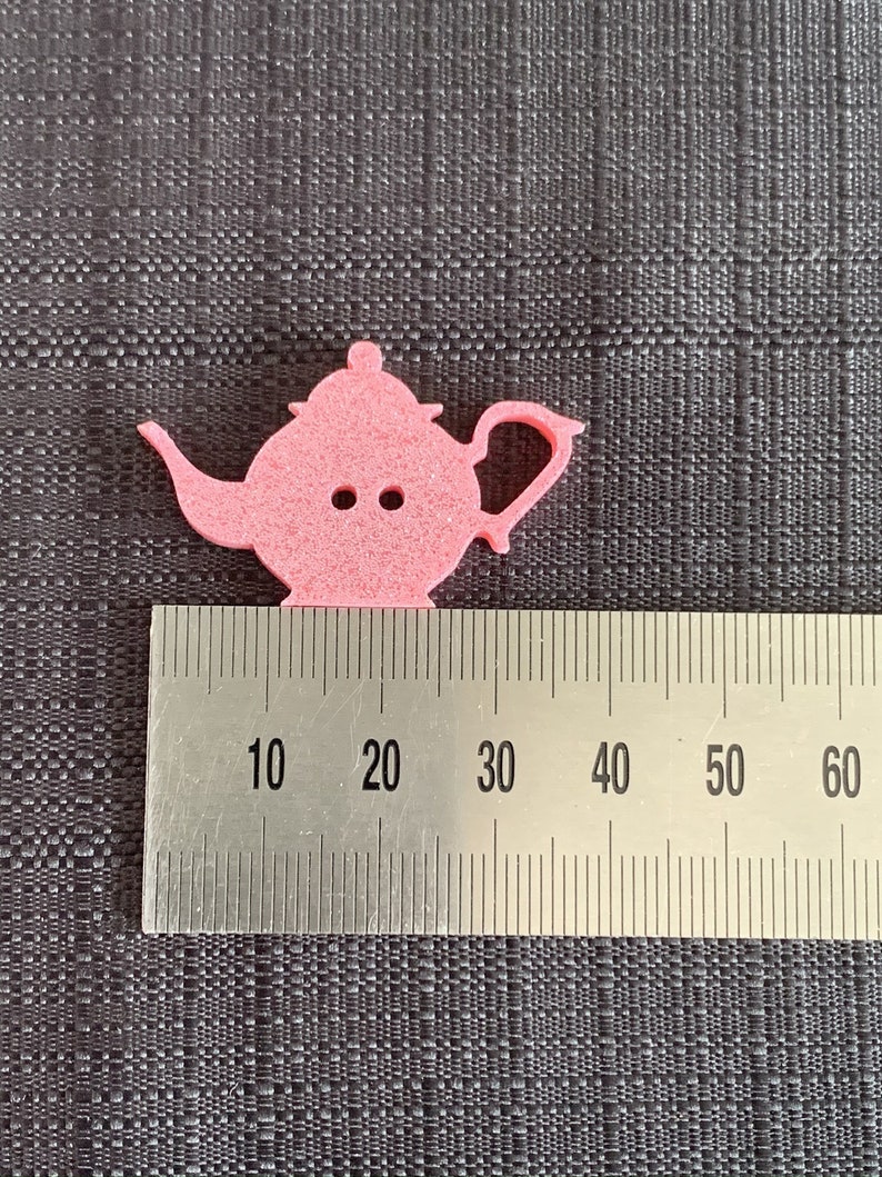 May include: A pink felt teapot cutout measuring approximately 2.5 inches wide. The teapot has a handle and a spout. The cutout is on a gray textured surface with a ruler for scale.