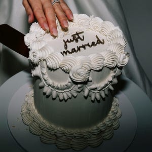 May include: A white tiered cake with piped frosting borders and a decorative ruffle design around the middle. The top of the cake features the text "just married" in dark lettering. A metal knife is positioned to slice into the cake.