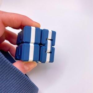May include: A blue and white fidget cube with a striped pattern. The cube is made of plastic and has a smooth surface. It is a popular toy for people of all ages.