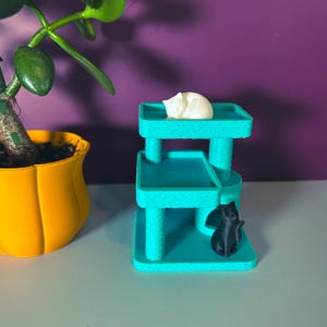 Miniature teal cat tree with textured scratching posts; black sitting cat figure and white sleeping cat on top platform; yellow planter and green plant for scale.