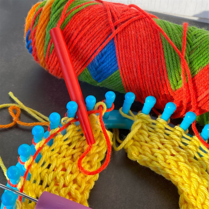 Red yarn feeder attached to striped skein of yarn resting on blue knitting loom with work in progress