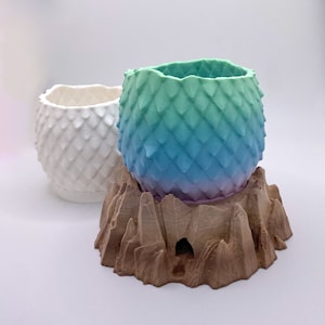 May include: Two dragon egg-shaped planters, one white and one with a gradient of green, blue, and purple, sitting on a brown, textured, tree stump-shaped base.