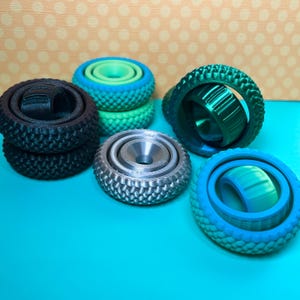 May include: A collection of colorful fidget spinners in various shades of blue, green, and silver. The spinners have a tire-like design with textured edges and a central bearing. Some are stacked, while others are displayed individually.
