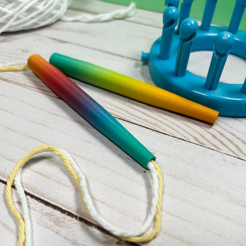 May include: Two tapered knitting tools with a smooth gradient finish, one transitioning from orange to red to blue and the other from green to yellow. A bright blue circular knitting loom with upright pegs sits nearby with white and yellow yarn.