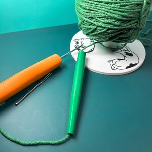 May include: A green yarn ball sits on a white circular disc featuring black cat illustrations. Beside it lies an orange crochet hook, a silver metal sewing needle, and a green cylindrical yarn guide tool with green yarn threaded through it.