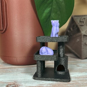 Miniature black cat tree with lavender sleeping and sitting cat figures, shown on wood surface with brown planter, green plant, and large dice decor in background